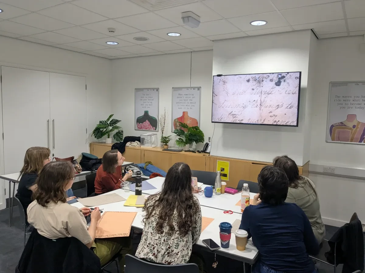Six researchers from the Queer History Club sit around a table covered in papers and coffee cups, and look up at a screen displaying a fragmented and damaged document from the Peglar Papers